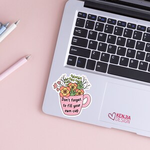 以下が含まれることがあります： ピンクのセラミック製のマグカップに花が飾られています。マグカップには持ち手があり、前面には「自分のカップを満たすことを忘れないで」という文字が印刷されています。