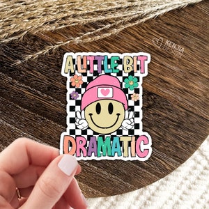 May include: A sticker featuring a smiley face wearing a pink beanie and the text "A little bit dramatic" in rainbow colours on a black and white chequered background.