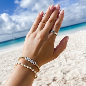 May include: A person's hand wearing two bracelets. The top bracelet is gold with white beads that spell out "SOPH". The bottom bracelet is gold with white pearl-like beads. The person is also wearing a silver ring on their ring finger.