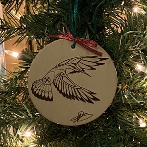 May include: A round, cream-coloured Christmas bauble featuring a brown and black stylised image of a bird with outstretched wings. The bauble has a green and red ribbon attached.