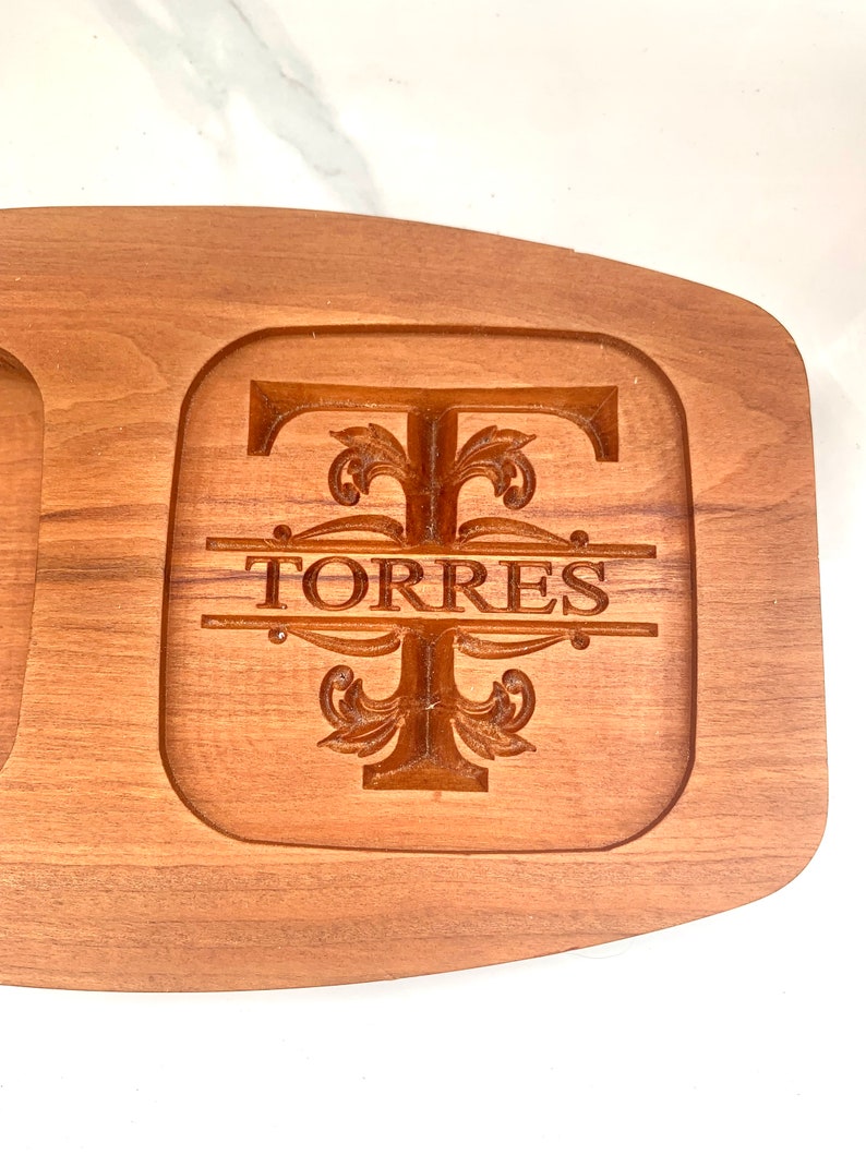 May include: A wooden serving tray featuring a carved design of the letter "T" with the name "TORRES" beneath. The tray has a natural wood finish and is ideal for serving snacks or appetizers.