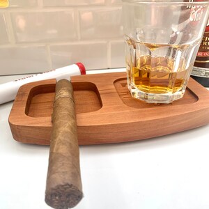 May include: A wooden tray with a cigar resting in a groove and a glass of amber liquid. The tray has a small, round indentation for an ashtray.