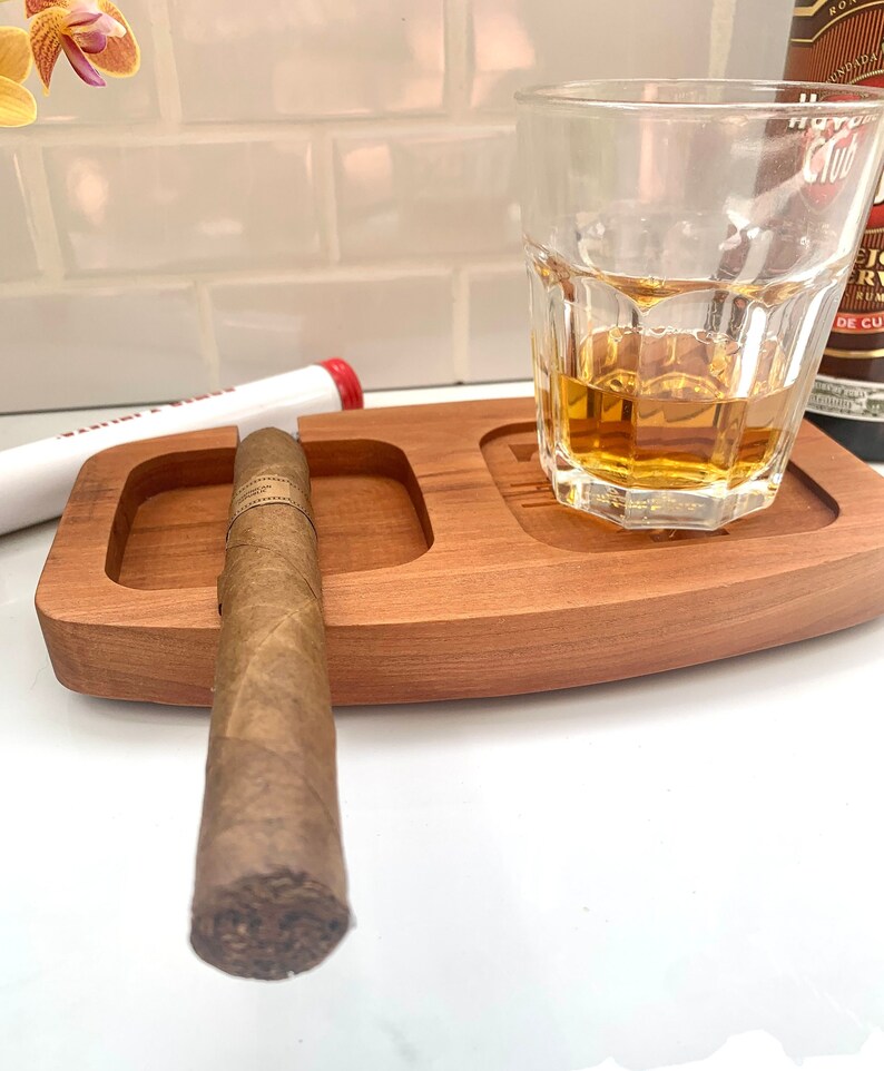 May include: A wooden tray with a cigar resting in a groove and a glass of amber liquid. The tray has a small, round indentation for an ashtray.