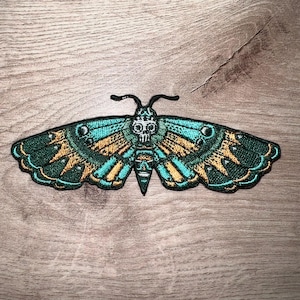 Puede incluir: Parche bordado con un diseño de polilla con una calavera en la cabeza. Las alas son de color turquesa y dorado con un borde verde. El parche está sobre una superficie de madera.