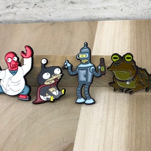 Puede incluir: Conjunto de cuatro pines esmaltados con personajes de dibujos animados. Incluye un alienígena rojo con bata blanca, una criatura pequeña con capa, un robot con una botella y una rana con collar. Contornos negros.