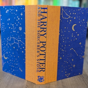 May include: A hardcover book titled "Harry Potter and the Sorcerer's Stone." The book's cover features a blue background with a constellation pattern and gold accents. The spine is orange with the title in gold lettering.