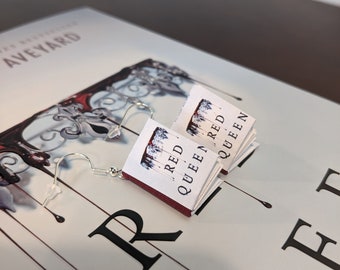 Op de afbeelding: Een paar zilveren boek oorbellen met een rode omslag en de tekst "Red Queen" op de voorkant. De oorbellen liggen op een wit oppervlak met het woord "Red" zichtbaar.