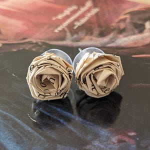 Puede incluir: Un par de pendientes de papel en forma de rosa de color crema. Las rosas están hechas de páginas de un libro y tienen texto negro impreso en ellas.