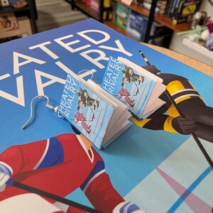 May include: A pair of handmade earrings featuring miniature book replicas with the title "Heated Rivalry." The earrings depict a hockey scene on a light blue background. The earrings are displayed on a blue surface with the book title visible.
