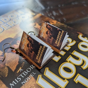 Puede incluir: Pendientes con forma de libro en miniatura, con la portada de "The Alloy of Law" de Brandon Sanderson. Los pendientes tienen pequeñas páginas y ganchos dorados, sobre un fondo de portada de libro.