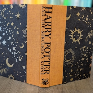 May include: A hardback book titled "Harry Potter and the Sorcerer's Stone" with a celestial design. The cover is black with gold moons, stars, and suns. The spine is orange with the title in gold lettering.