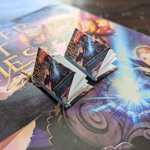 May include: A pair of book-shaped earrings featuring the cover of the book "Keeper of the Lost Cities" by Shannon Messenger. The earrings are made of paper and have gold-colored hooks.