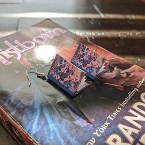 Puede incluir: Un par de pendientes en forma de libro con una cubierta azul y la palabra "Mistborn" impresa en la parte delantera. Los pendientes cuelgan de ganchos plateados.