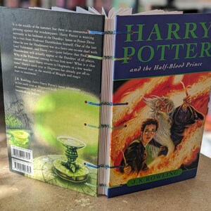 May include: A hardcover book titled "Harry Potter and the Half-Blood Prince" is open, revealing the spine and back cover. The cover features Harry, Dumbledore, and fiery imagery. The spine is hand-stitched with blue and green thread. The back cover has text.