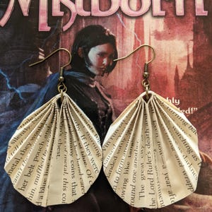 Puede incluir: Un par de pendientes de páginas de libro con fondo blanco y ganchos dorados. Los pendientes tienen forma de abanico y están hechos de páginas de un libro. El texto en las páginas es visible y dice "Mistborn" y "New York Times bestselling author".