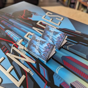 Puede incluir: Un par de pendientes de plata con abalorios de libros en miniatura. Los libros son azules y negros con el título "Renegades" impreso en la portada.