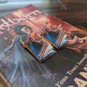 Puede incluir: Un par de pendientes en forma de libro con una cubierta azul y morada y detalles dorados. Los pendientes muestran el título "Mistborn" y el autor "Brandon Sanderson".