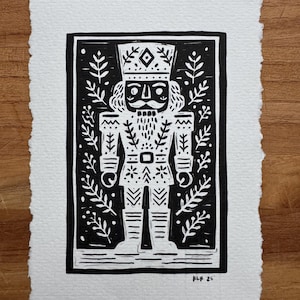 May include: A black and white linocut print of a nutcracker soldier. The print features a detailed design of the nutcracker with a mustache, crown, and decorative uniform. The background includes leaf-like patterns. The print is on textured white paper.