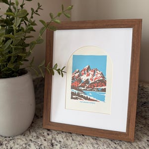 Pu&ograve; includere: Una stampa incorniciata raffigurante un paesaggio montano con cime rosse e bianche, un lago blu e alberi verdi. La stampa &egrave; in una cornice di legno marrone con un passe-partout bianco. Il testo "THE GRAND TETON" &egrave; in basso.