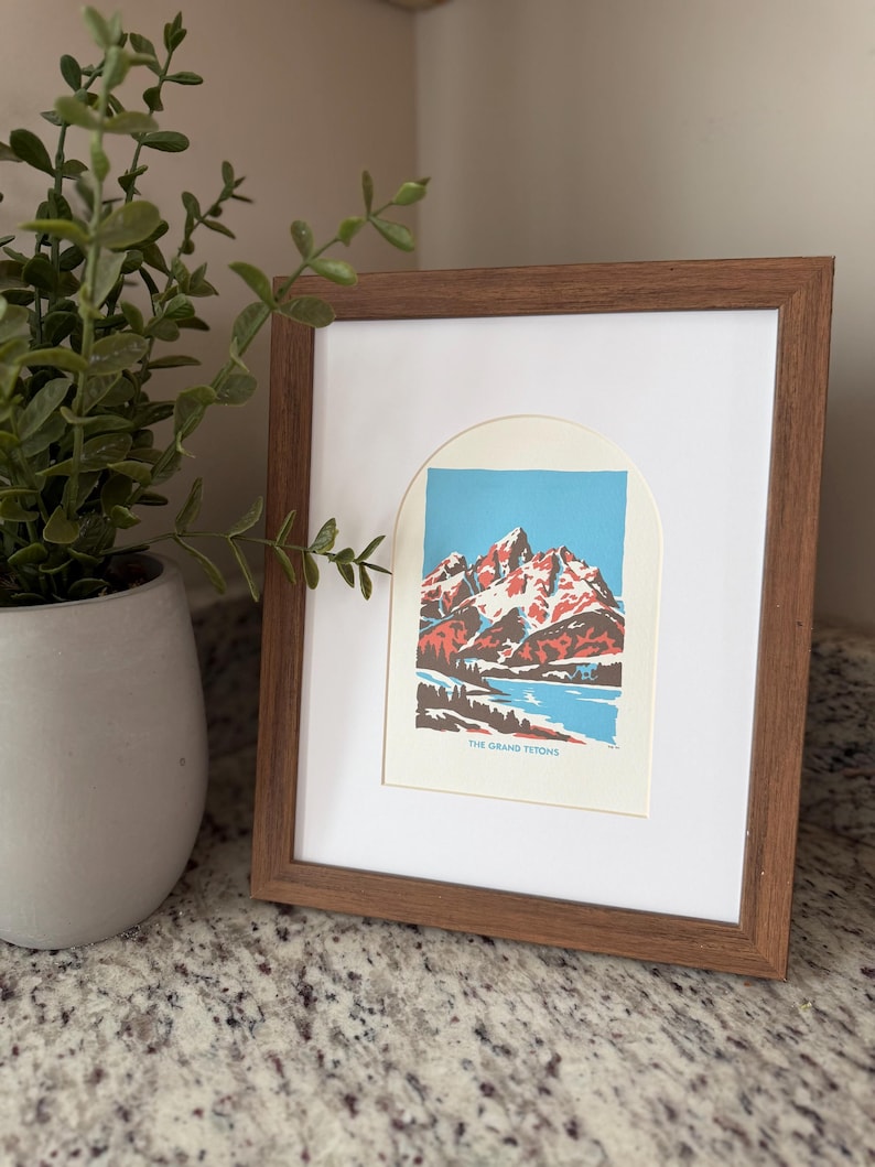 Pu&ograve; includere: Una stampa incorniciata raffigurante un paesaggio montano con cime rosse e bianche, un lago blu e alberi verdi. La stampa &egrave; in una cornice di legno marrone con un passe-partout bianco. Il testo "THE GRAND TETON" &egrave; in basso.