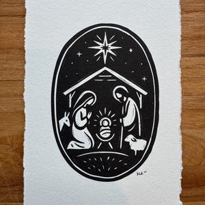May include: Black and white linocut print depicting the Nativity scene within an oval frame. The image features Mary and Joseph kneeling before the baby Jesus, with a star above and a sheep nearby. The artwork is on textured paper.