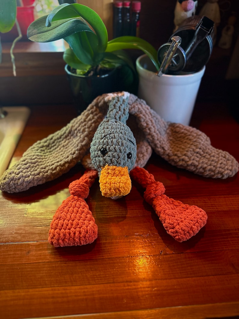 May include: A crocheted gray duck toy with orange feet and a yellow beak. The duck is lying on a wooden surface.