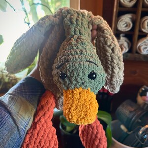 May include: A crocheted plush toy duck with a green body, orange beak, and orange feet. The duck is hanging upside down.
