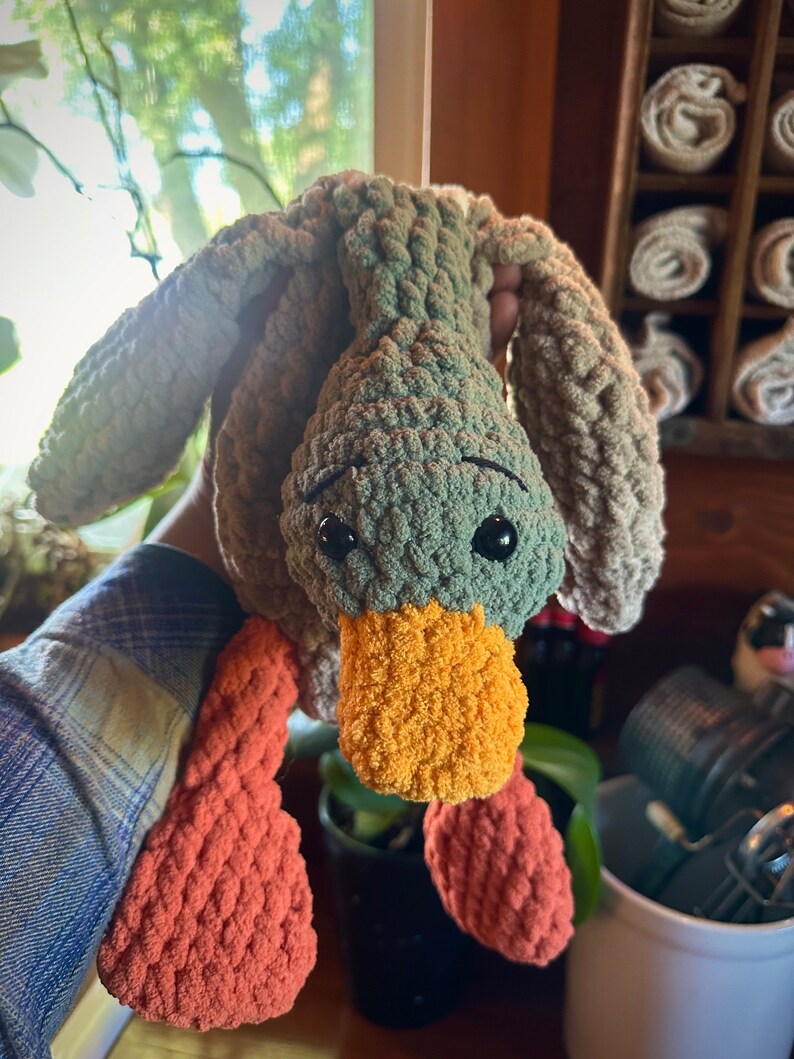 May include: A crocheted plush toy duck with a green body, orange beak, and orange feet. The duck is hanging upside down.