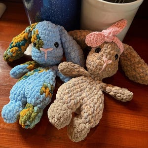 May include: Three handmade crochet bunny plushies in blue, tan, and tan with a pink bow. The bunnies have black button eyes and pink embroidered noses. The ears and feet have a textured design.