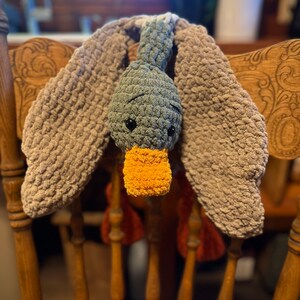 May include: A crocheted gray duck with a yellow beak and brown wings hanging on a wooden chair.
