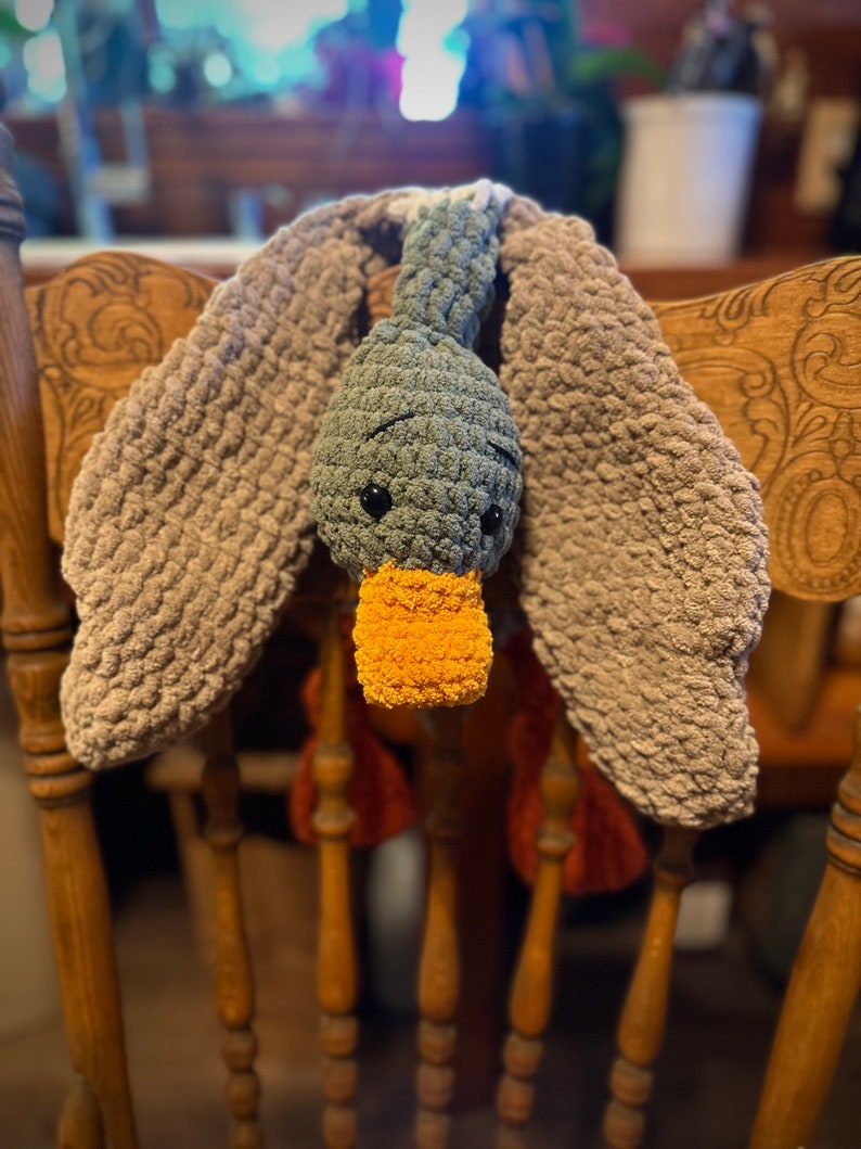 May include: A crocheted gray duck with a yellow beak and brown wings hanging on a wooden chair.