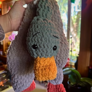 May include: A crocheted plush toy shaped like a duck with a gray head, orange beak, and pink feet. The duck is hanging from a hand.