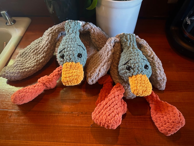 May include: Two crocheted plush duck toys with orange and gray bodies and blue heads. The ducks have orange beaks and black eyes.