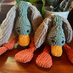 May include: Two crocheted plush duck toys with gray and orange fabric. The ducks have large eyes and a yellow beak.
