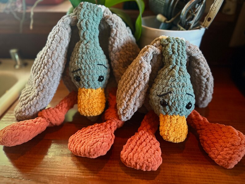 May include: Two crocheted plush duck toys with gray and orange fabric. The ducks have large eyes and a yellow beak.