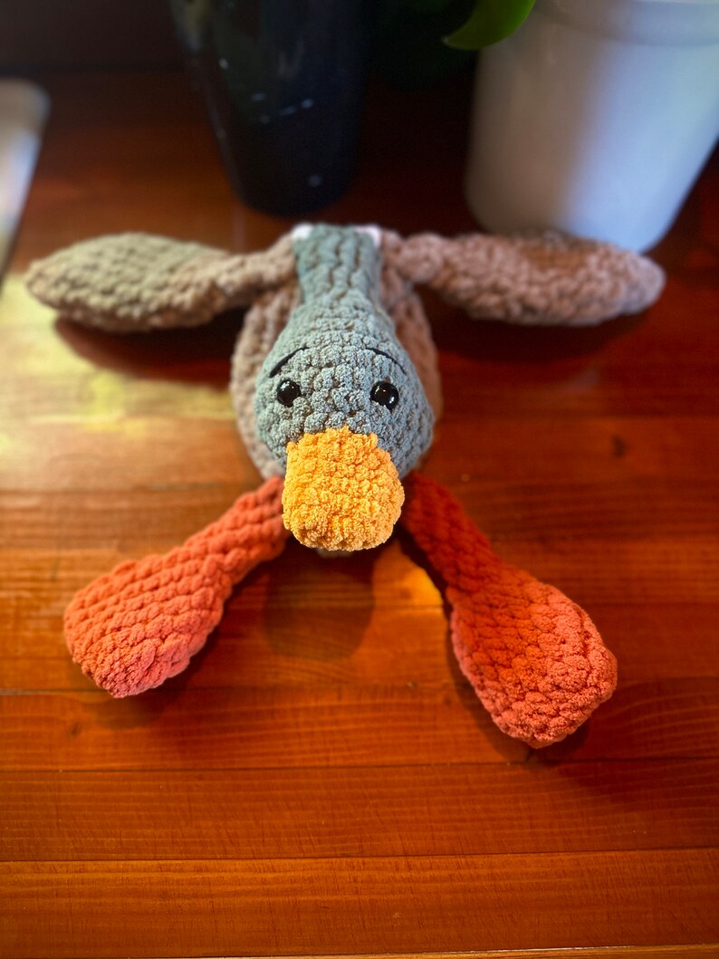May include: A crocheted stuffed duck toy with brown, gray, and orange yarn. The duck has a long orange beak and black eyes.