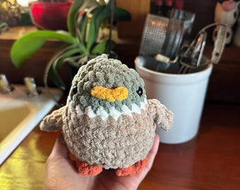 May include: A small, crocheted bird plush toy with a green, brown, and orange color scheme. The bird has a yellow beak and black eyes.