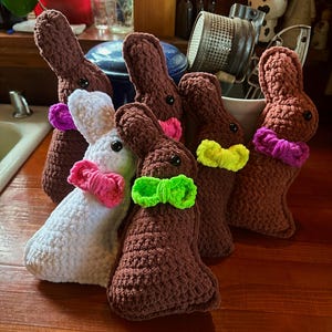 May include: A group of six crocheted bunny plushes, each with a different colored bow. The bunnies are brown and white, and the bows are pink, green, and yellow.