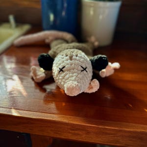 May include: A crocheted stuffed animal resembling an opossum with black eyes and a white and brown body. The opossum is lying on a wooden surface.
