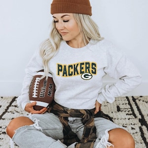 May include: A light gray sweatshirt with the word "PACKERS" in green and gold lettering, and a green "G" logo. The person is holding a brown football. A brown beanie and ripped jeans complete the look.