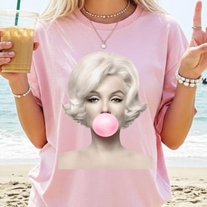 May include: Pink t-shirt featuring a graphic of Marilyn Monroe blowing a bubblegum bubble. The image is a black and white portrait with a pink bubble. The background is a beach scene.