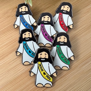 May include: A collection of enamel pins featuring a cartoon depiction of Jesus. Each pin has a white robe, black hair, and a sash with the words "Jesus ❤️ You" in various colors, including blue, red, purple, green, and yellow.
