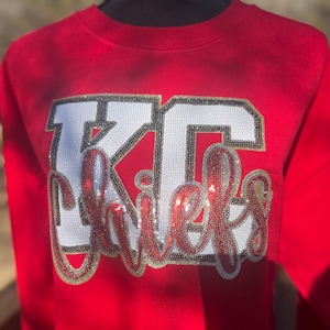 May include: A red sweatshirt with the text "KC Chiefs" in white, red, and gold sequins. The letters "KC" are in a block style, and "Chiefs" is in a cursive font. The sweatshirt is displayed on a black mannequin.
