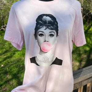 May include: A pink t-shirt with a black and white image of Audrey Hepburn blowing a bubble. The bubble is pink.