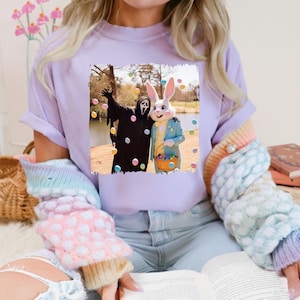 May include: A light purple t-shirt featuring a square image of the Ghostface killer from the Scream movies and the Easter Bunny posing together. The Easter Bunny is holding a basket of colorful eggs. The shirt is a casual wear item.