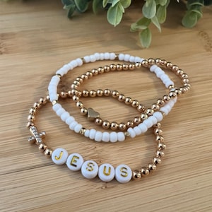 Peut inclure: Trois bracelets de perles. L'un des bracelets comporte le mot "JESUS" épelé en perles de lettres blanches. Les autres bracelets sont perlés d'or et de blanc. Un bracelet a un petit cœur doré et un autre une breloque en forme de croix.