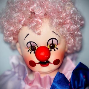 May include: A vintage clown doll with a painted face, red nose, and pink curly wig. The doll wears a pink and lavender ruffled collar and a blue bow with red polka dots. The clown has painted rosy cheeks and a sad expression.