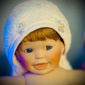 May include: A porcelain doll with blue eyes and blonde hair, wearing a white terry cloth bonnet adorned with clear, iridescent beads. The doll has rosy cheeks and a gentle smile, creating a serene and innocent expression.