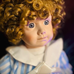 May include: A vintage porcelain doll with curly blonde hair and blue eyes. The doll is wearing a blue and white striped dress with a white collar and bow. The doll's face has a serene expression.
