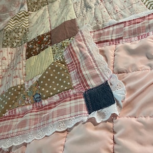 May include: A patchwork quilt featuring a variety of fabric squares in shades of pink, brown, white, and tan with floral, polka dot, chevron, and plaid patterns. The quilt has a white eyelet lace trim border and a solid light pink backing.